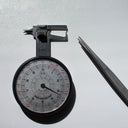 3.3 mm round brilliant cut diamond held in an analog millimeter gauge beside a pair of jeweler’s tweezers on a white surface.