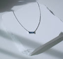 Bar None baguette sapphire necklace in 14K white gold displayed on a white stone slab with dramatic shadows.