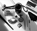 Black-and-white view of a jewelry designer’s desk with diamonds, a loupe, ring sizers, gauges, tweezers, a calculator displaying 777, tea, and a scale weighing a 7.077-carat stone.